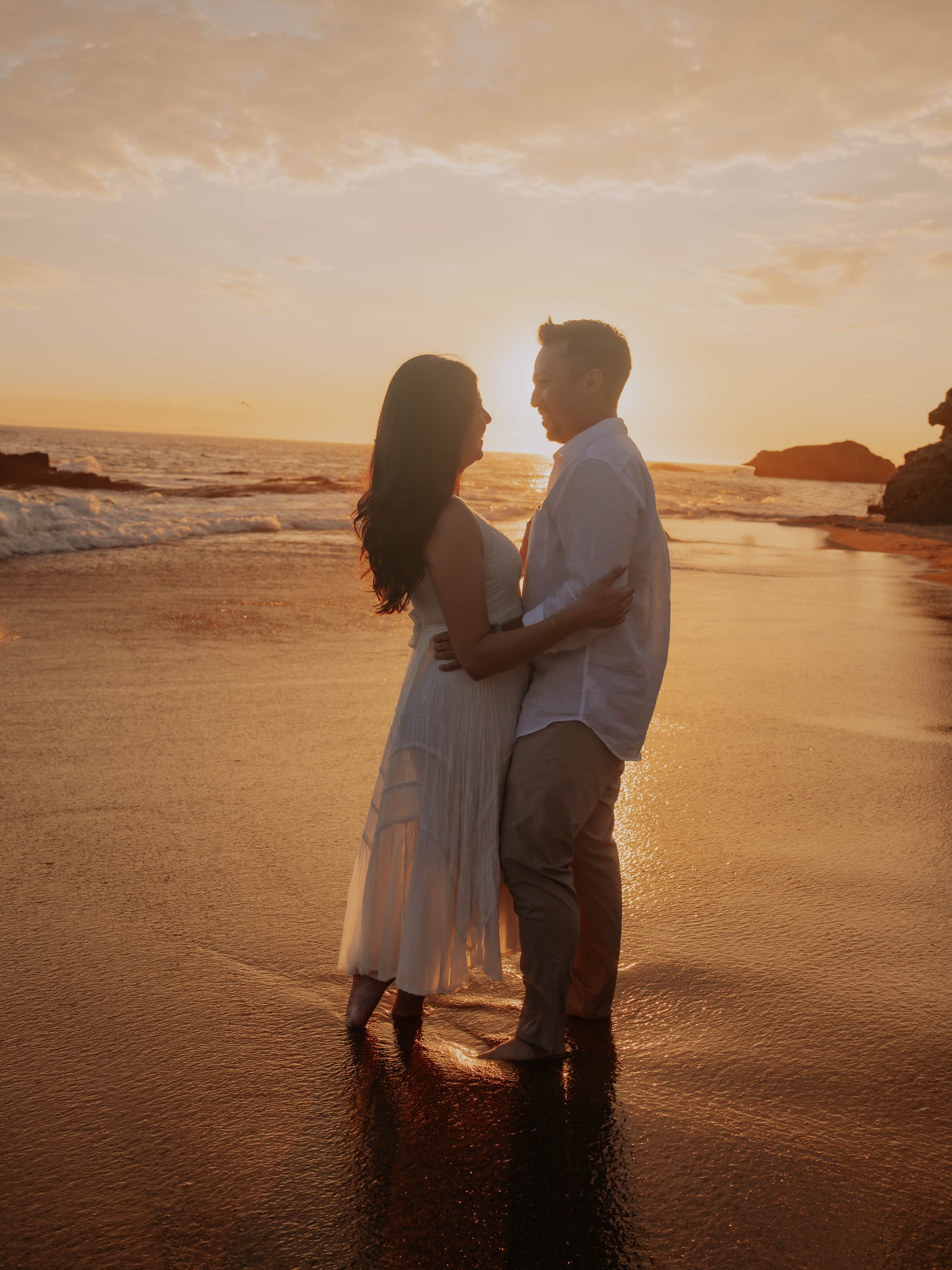 Guadalupe and Harrison embracing on the beach at sunset, silhouetted against the glowing sky