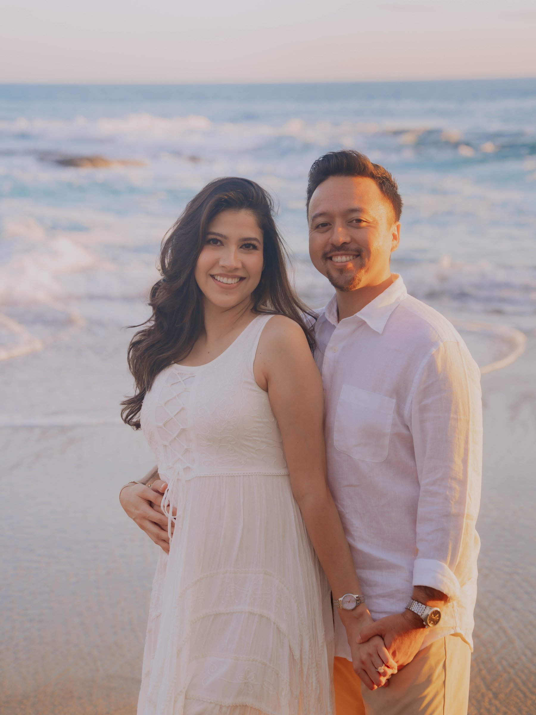 Guadalupe and Harrison embracing on the rocks as a wave crashes behind them