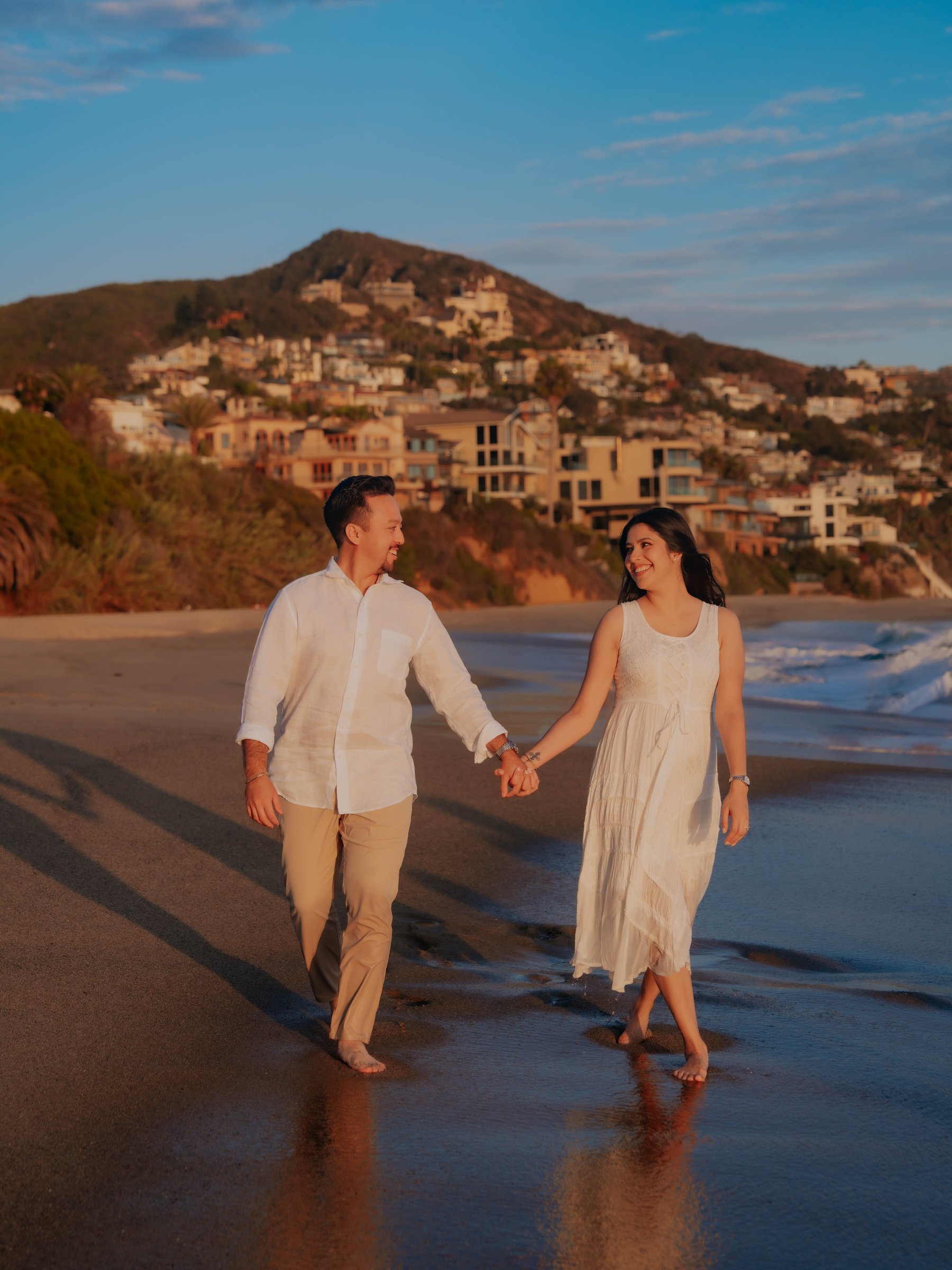 Guadalupe and Harrison walking hand in hand along the beach at sunset, a hillside town in the background