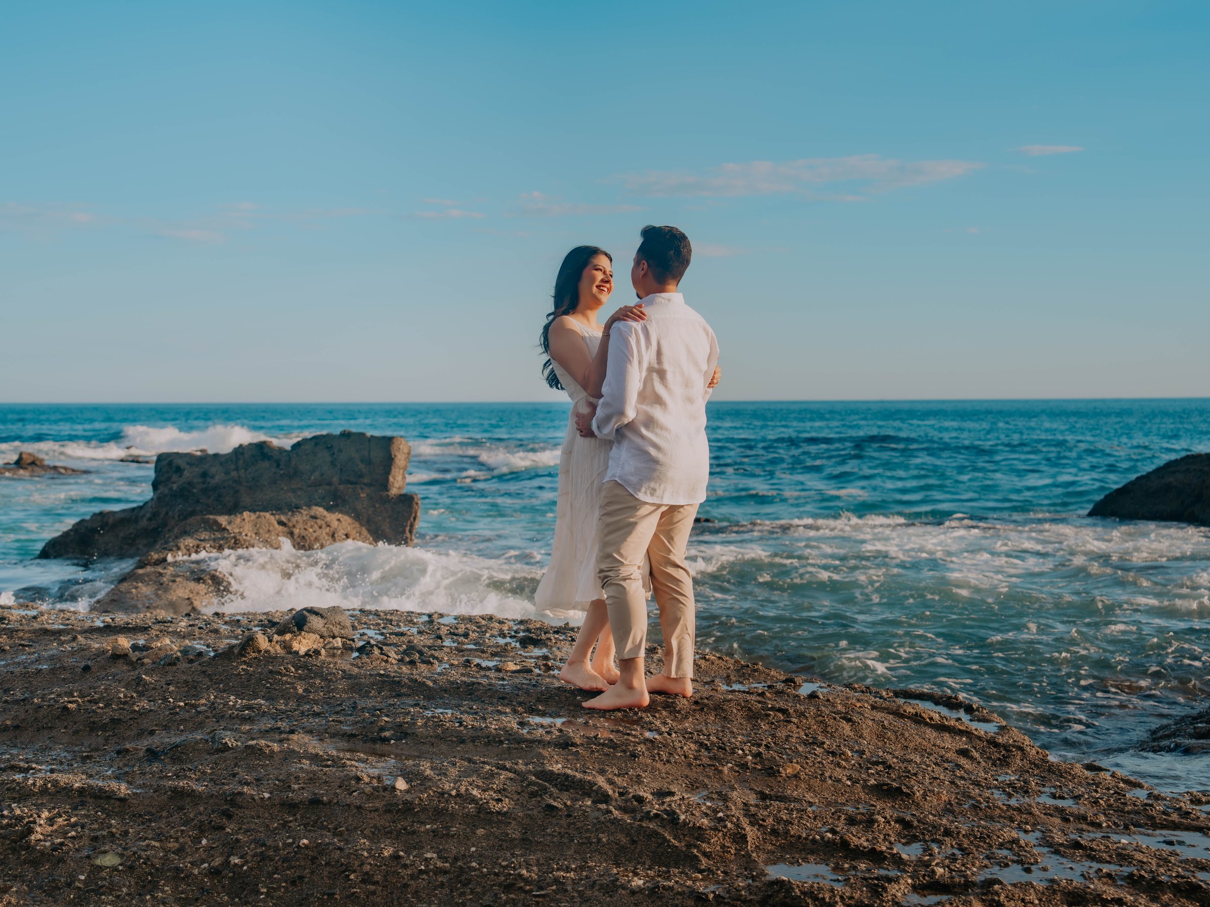 Guadalupe and Harrison embracing on the rocks by the ocean under a clear blue sky